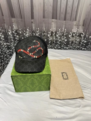 Svart Gucci keps med ormtryck - Snygg svart keps från Gucci med klassiskt GG-mönster och en röd-vit orm på framsidan. Kepsen har böjd skärm och justerbar rem baktill. Materialet är canvas med detaljer i mesh på sidorna för extra komfort.Äkta Keps kom privat om du är intresserad 