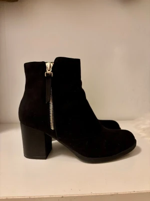 Svarta mocka stövletter med klack - Snygga svarta stövletter i mocka med grov klack och dragkedja i silver på sidan. Klassisk rund tå och stilren design som passar till många outfits. Perfekta för dig som vill ha en enkel men trendig look.