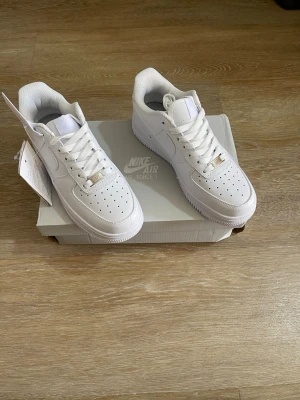 Nike Air Force 1 vita sneakers - Klassiska Nike Air Force 1 sneakers i helvitt läder med perforerad tå, vit sula och vita skosnören. Ikonisk Nike Swoosh på sidorna och metallbricka på snörningen. Perfekt för en clean och stilren look.