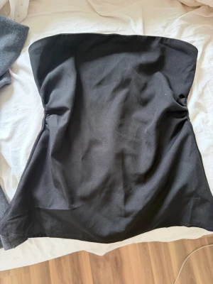 Svart axelbandslös tubtopp - En stilren svart tubtopp. Axelbandslös design som sitter tight över bysten och har en enkel, klassisk form. Perfekt att styla med jeans eller kjol för en trendig look.
