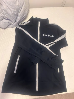  Palm Angels - Säljer en svart zip hoodie från Palm Angels med vita detaljer längs ärmar och dragkedjor. Hoodien har hög krage, långa ärmar och två fickor med dragkedja. Materialet är mjukt och sportigt, perfekt för en cool och avslappnad look bra skick inga fel med den stolek xs pris 200 kr 