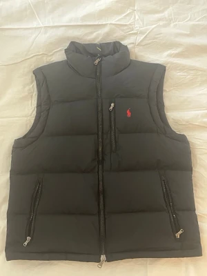 Svart dunväst från Polo Ralph Lauren - Svart dunväst från Polo Ralph Lauren med hög krage, två sidofickor med dragkedja och klassisk röd logotyp på bröstet. Västen har en rak passform och är tillverkad i ett quiltat material som håller värmen. Perfekt för lager-på-lager och snygg till jeans.