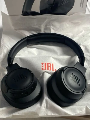 JBL svarta over-ear hörlurar - Snygga svarta over-ear hörlurar från JBL med robust design och mjuka kuddar för extra komfort. Hörlurarna har en stilren matt finish och integrerade knappar på sidan för enkel styrning. Perfekt för dig som vill ha kraftfullt ljud och cool look.