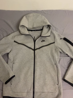 Grå Nike hoodie med dragkedja - Snygg grå hoodie från Nike med hel dragkedja och svarta detaljer. Hoodien har en klassisk huva, svarta linjer över bröstet och Nike-logga på vänster sida. Tillverkad i mjukt sweatshirtmaterial, perfekt för en sportig och avslappnad stil. Den passar för både M och S. Pris kan diskuteras så hör av dig vid minsta intresse. 