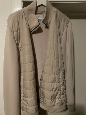 Beige pufferjacka med ribbad krage, L från Bläck - Stilren beige pufferjacka med quiltad front och ribbad krage. Jackan har släta ärmar och baksida i mjukt material, samt ribbade muddar vid ärmslut och nederkant. Perfekt för kalla dagar och enkel att matcha med olika outfits.