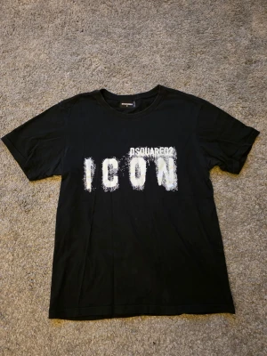 Svart DSQUARED2 ICON t-shirt - Svart t-shirt från DSQUARED2 med stort vitt ICON-tryck på bröstet och DSQUARED2-logga ovanför. Klassisk passform och rund halsringning. Materialet är mjuk bomull och trycket har en distressed look.