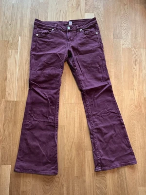 Burgundy  bootcut jeans med fickor - Snygga burgundy jeans med bootcut passform och klassiska femficksdetaljer. Jeansen har två bakfickor med lock och silverknappar samt raka ben med utsvängd nederkant. Materialet är mjukt och slitstarkt, perfekt för dig som vill ha en cool look. 