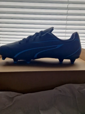 Svarta och blåa Puma fotbollsskor - Snygga fotbollsskor från Puma i svart med blå detaljer och logga. Skorna har klassisk låg profil, snörning och fasta dobbar i plast för spel på gräs. Tillverkade i slitstarkt syntetmaterial som ger bra bollkontroll och grepp på planen.