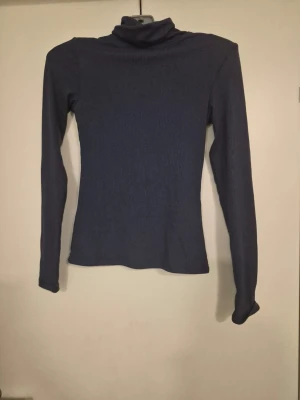 Mörkblå ribbad polotröja - En mörkblå långärmad polotröja med ribbad struktur och figurnära passform. Tröjan har hög krage och är tillverkad i ett stretchigt material som sitter snyggt på kroppen. Perfekt för lager-på-lager och stilrena outfits.