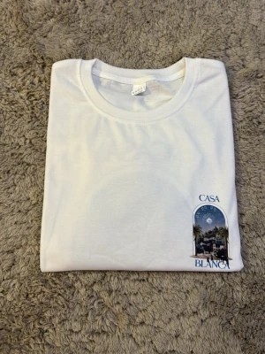  Casa Blanca t-shirt  - Snygg vit t-shirt från Casa Blanca med ett litet tryck framtill som visar en stadsvy med palmer och blå himmel. T-shirten har klassisk rund hals och korta ärmar. Perfekt för dig som gillar stilrena plagg med en twist.