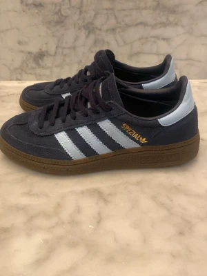 Adidas Spezial sneakers mörkblå - Säljer ett par Adidas Spezial sneakers i mörkblå mocka med ljusblå detaljer och klassiska tre ränder på sidan. Skorna har bruna gummisulor och guld Spezial-logga på sidan. Perfekt för dig som gillar retrostil och ikoniska streetwear-vibes.