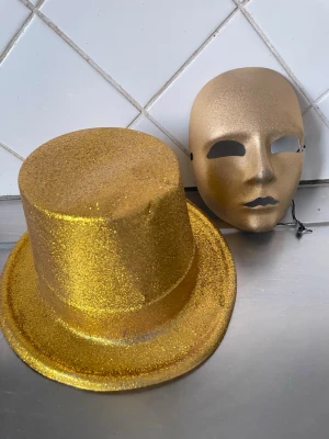 Glittrig guldhatt & Guld Mask som i förrädarna  - Säljer en glittrig guldhatt i plast och en enkel Guld  mask  samma som förrädarna har med elastiskt band. Perfekt för maskerad, temafest eller cosplay. Hatten har hög form och masken täcker hela ansiktet.samma mask som Förädarna på Tv har 