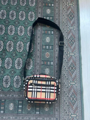 Burberry axelväska med rutigt mönster - Säljer en snygg axelväska från Burberry med det klassiska beige, svarta, vita och röda rutmönstret. Väskan har justerbar svart axelrem, dragkedja framtill och detaljer i silver. Perfekt för dig som gillar stilrena accessoarer med lyxig känsla.