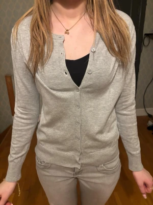 Ljusgrå klassisk kofta - En stilren ljusgrå kofta med rund halsringning och knäppning framtill. Koftan har långa ärmar och är tillverkad i ett mjukt, tunt stickat material. Perfekt att slänga över en topp för en enkel och clean look.