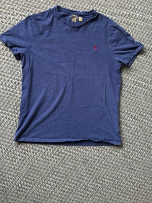 Blå t-shirt från Polo Ralph Lauren - Klassisk blå t-shirt från Polo Ralph Lauren med korta ärmar och rund hals. Liten röd broderad logga på bröstet. Skön och enkel modell i mjuk bomull. Funkar i Medium också