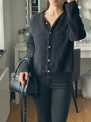 Kofta med guldknappar från H&M - Stilren svart kofta med guldiga knappar framtill och två dekorativa fickor på bröstet. Koftan är stickad i ett grovt mönster och har en rak, något boxig passform med långa ärmar. Perfekt för lager-på-lager och enkel att matcha med olika outfits.