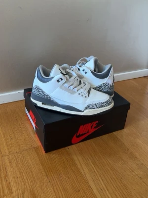 Nike Air Jordan 3 White Cement - Nike Air Jordan 3 White Cement sneakers med vit läderöverdel, grå detaljer och ikoniskt elefantmönster på tå och häl. Skorna har svarta och grå accenter, klassisk Jumpman-logga på plös och häl samt en robust sula. Perfekta för dig som gillar streetwear och retrostil.