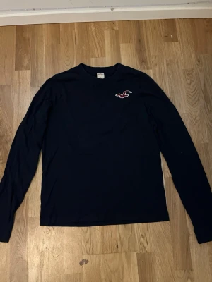 Mörkblå långärmad topp Hollister XL - Mörkblå långärmad topp från Hollister med klassisk logga broderad på bröstet. Enkel och stilren design med rund halsringning, perfekt för en avslappnad look. Tillverkad i mjukt material som känns skönt mot huden.