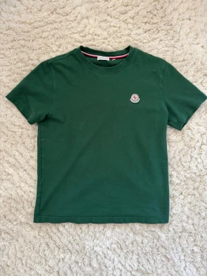 Grön Moncler t-shirt  - Snygg grön t-shirt från Moncler med klassisk rund hals och korta ärmar. På bröstet sitter Monclers ikoniska logotyp i vitt, rött och blått. T-shirten har en diskret röd, vit och blå detalj på insidan av kragen. Tillverkad i mjuk bomull. Hör av dig om du vill diskutera pris!