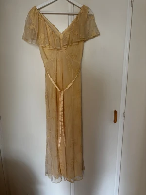 Gul spetsklänning från Newbie XS - Romantisk gul klänning från Newbie i storlek XS. Klänningen har offshoulder-modell, volang upptill och är dekorerad med broderade blommor i spets. Lång och luftig passform med knytband i midjan. Perfekt för sommarens alla tillfällen. Använd 1 gång
