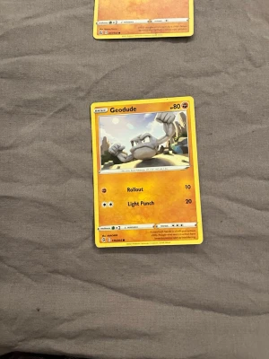 Pokémonkort Geodude - Pokémonkort med Geodude-motiv. Kortet har en gul-orange ram och visar Geodude i en tecknad miljö. Text och symboler är tryckta på framsidan, och kortet är gjort av laminerad kartong. Perfekt för samlare eller Pokémon-fans.