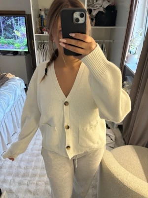 KOFTA - Mysig vit kofta med djup V-ringning, stora bruna knappar och två framfickor. Koftan har en avslappnad passform och ribbstickade muddar vid ärmslut och nederkant. Perfekt att slänga på sig över en t-shirt eller linne.