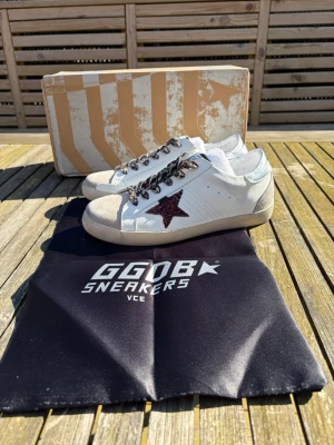 Golden goose vita sneakers med stjärna - Snygga vita sneakers från Golden Goose med en glittrig mörkröd stjärna på sidan, leopardmönstrade skosnören och grå mockadetaljer på tån. Skorna har en klassisk låg modell och slitna detaljer för en cool look. Perfekta för dig som vill sticka ut.