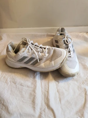 Vita Adidas sneakers med mesh och ränder - Säljer ett par vita Adidas sneakers med klassiska tre grå ränder på sidan. Yttersulan är mönstrad för extra grepp och skorna har lågt skaft. Perfekta för en sportig look. Något slitna och påväg till hål. Men fungerar fortfarande fint ännu... 39 och 1/3