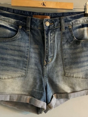 Blå jeansshorts från Gina Tricot - Snygga blå jeansshorts från Gina Tricot med klassisk femficksdesign och uppvikta benslut. Shortsen har en normal passform och är tillverkade i bomull. Perfekta för sommaren och enkla att matcha med olika toppar.