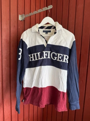Randig Hilfiger rugbytröja - Säljer en klassisk långärmad rugbytröja från Tommy Hilfiger med breda ränder i vitt, marinblått och vinrött. Tröjan har stor logga framtill, nummerdetalj på ärmen och krage. Tillverkad i mjuk bomull och har knappar vid halsen.