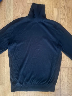Svart polotröja från Paul Smith - Ny helt oanvänd. Klassisk svart polotröja från Paul Smith i mjukt material. Tröjan har lång ärm, hög polokrage och ribbade muddar vid ärmslut och nederkant. Perfekt för en stilren och enkel look.