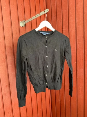 Svart kofta från Ralph Lauren Sport - Klassisk svart kofta i 100% merinoull från Ralph Lauren Sport. Rund halsringning, knäppning framtill med vita knappar och diskret broderad logga på bröstet. Perfekt att slänga över en t-shirt eller klänning för en stilren look. OBS! BARN STORLEK XL, passar xs/S Dam