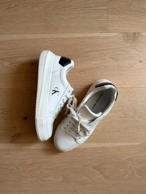 Vita Calvin Klein sneakers - Endast anvönda två gånger! Snygga vita sneakers från Calvin Klein med svart detalj på hälen och klassisk CK-logga på sidan. Skorna har låga skaft, vita snören och en stilren design i slätt lädermaterial. Perfekta för en clean och modern look.