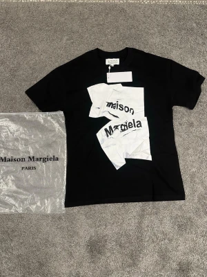 Maison Margiela svart t-shirt - Maison Margiela t-shirt i svart med unikt vitt tryck på framsidan där loggan är splittrad över flera fält. Klassisk rund hals och korta ärmar. Materialet är mjuk bomull och medföljande dustbag ingår.