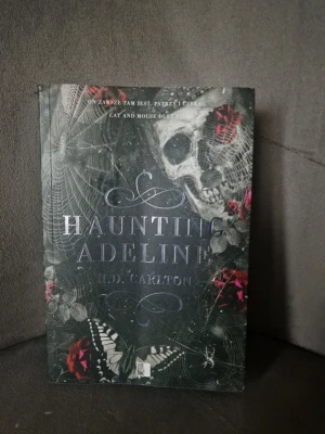Haunting Adeline av H.D. Carlton - Säljer boken 'Haunting Adeline' av H.D. Carlton. Omslaget är svart med detaljer av spindelnät, dödskalle, röda rosor och fjärilar vilket ger en mörk och mystisk känsla. Perfekt för dig som gillar spänning och skräckromaner. den är på POLSKA 
