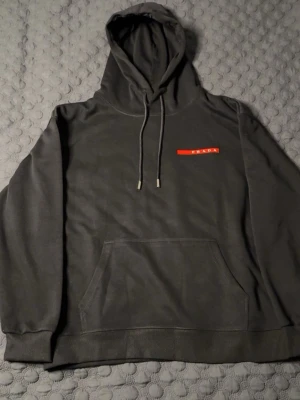Svart hoodie från Prada - Svart hoodie från Prada med klassisk huva och dragsko. På bröstet sitter en röd logga med vit text. Hoodien har en stor magficka och ribbade muddar vid ärmslut och nederkant. Perfekt för en stilren och trendig look. STORLEK M, PASSAR M. HELT NY, PRISLAPP PÅ