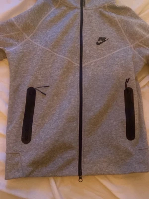  Nike tech hoodie  - Snygg grå hoodie från Nike med svarta dragkedjor och logga på bröstet. Tröjan har hel dragkedja framtill, två stora fickor med svarta dragkedjor och en skön huva. Perfekt för en sportig och avslappnad stil.