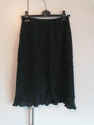 Prickig midikjol från danskt märke i viskos  - Svart midikjol med vita prickar och volangkant nertill. Kjolen har en rak passform och är tillverkad i ett lätt och mjukt tyg som ger ett fint fall. Perfekt för dig som gillar klassiska mönster med en söt twist. Gjord i viskos. 