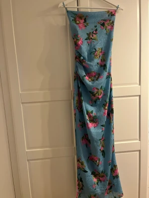 Blommig blå långklänning med snörning från Adoore - Långklänning i ljusblått med blommigt mönster i rosa och grönt. Klänningen har smala axelband och snörning i ryggen för en snygg detalj. Tillverkad i ett lätt, glansigt material som ger en somrig känsla.