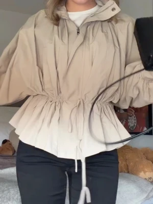 Beige jacka med snörning i midjan - Trendig beige jacka med hög krage och dragkedja framtill. Jackan har snörning i midjan för en snygg siluett och vida ärmar med resår vid handlederna. Perfekt för lager-på-lager och en chill streetstyle-look. 