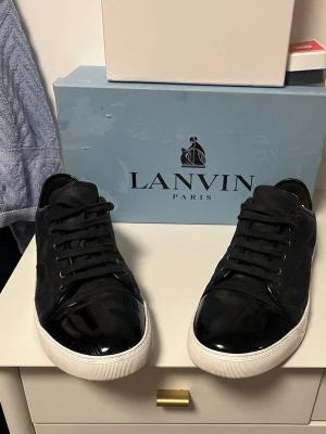 Svarta Lanvin sneakers med lackdetalj - Snygga svarta sneakers från Lanvin med glansig lackad tå och vit sula. Skorna har klassisk låg profil, svarta skosnören och stilren design. Perfekta för dig som gillar exklusiva och trendiga sneakers med en unik touch.