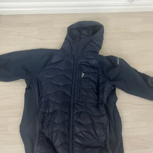 Svart vindjacka med huva från Peak Performance - Snygg svart vindjacka från Peak Performance med quiltade partier framtill och på ryggen. Jackan har huva, hel dragkedja och diskret logga på bröstet. Ärmar och sidor är i stretchigt material för extra komfort och rörelsefrihet.