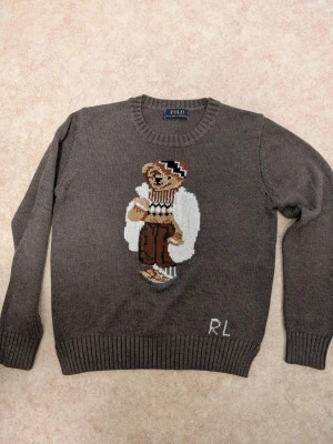 Brun/mörkgrön stickad tröja Polo Ralph Lauren - Stickad brun tröja från Polo Ralph Lauren med ikonisk björn på bröstet. Storlek L men passar som S/M. Tröjan har rund halsringning, ribbade muddar och RL-broderi nedtill. Perfekt för dig som gillar klassiska och lekfulla detaljer.