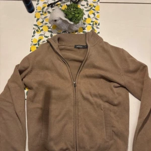 Beige ziptröja med ribbad krage - Säljer en beige ziptröja från dressman  med ribbad ståkrage och två fickor framtill. Tröjan har långa ärmar och ribbade muddar vid ärmslut och nederkant. Perfekt för lager på lager och enkel att matcha med olika outfits.