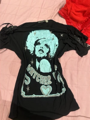 Svart t-shirt med turkost tryck Watcher - Cool svart t-shirt från Watcher med stort turkost tryck av ett ansikte och texten 'Watcher' framtill. T-shirten har korta ärmar med snörningsdetaljer och en avslappnad passform. Perfekt för dig som gillar unika statement-plagg.