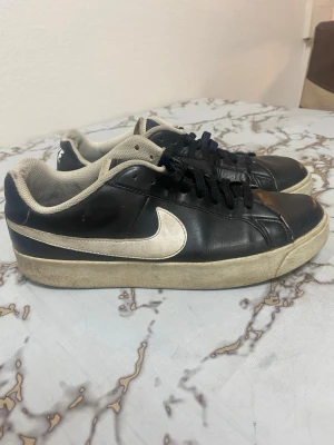 Nike Royale Low svarta sneakers - Svarta Nike Royale Low sneakers i klassisk modell med vit swoosh på sidan och beige detaljer. Skorna har svart ovandel i läder, svart snörning och beige innersula samt yttersula. Perfekta för dig som gillar en retro och stilren look. Fina skor behöver kanske bara liten tvätt