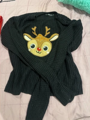 Svart stickad tröja med renmotiv - Mysig svart stickad tröja med ett gulligt renmotiv framtill. Tröjan har rund halsringning och långa ärmar med ribbade muddar. Perfekt för vintern och julen när du vill ha något både varmt och lekfullt.