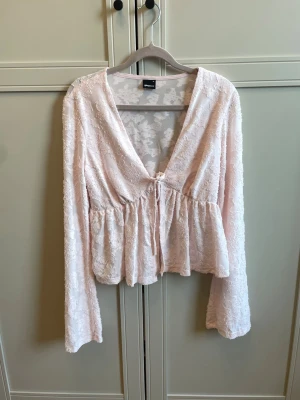 Ljusrosa spetsblus med knyt - Supersöt ljusrosa blus från Gina i spets med v-ringning och knyt framtill. Blusen har långa ärmar och volang nedtill för en romantisk vibe. Perfekt att styla med jeans eller kjol för en trendig look. Använd i max 2h.