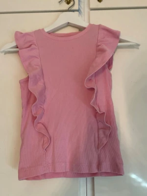 Rosa ribbad topp  - Supersöt ljusrosa ribbad topp från Kappahl med volangdetaljer längs axlar och sidor. Ärmlös modell som är perfekt till sommaren och har en mjuk, stretchig känsla. Enkel att matcha med jeans eller kjol för en trendig look.