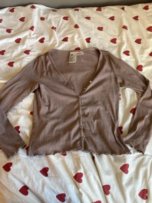 Brun ribbad kofta från H&M - Säljer en tunn, brun ribbad kofta från H&M med v-ringning och långa ärmar. Koftan har små vita knappar framtill och vågig kant längst ner. Perfekt att slänga över en topp för en chill look.
